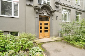Un edificio con una puerta de madera grande, un porche pequeño y un jardín con diversas plantas.
