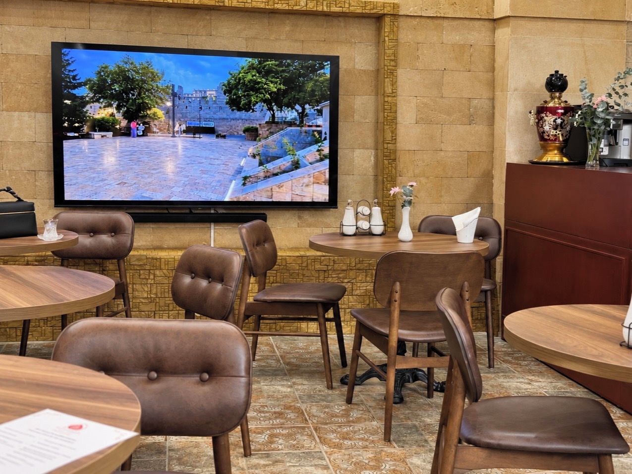 Ein Restaurantinneres mit runden Tischen, braunen Ledersesseln, einem Flachbildfernseher an der Wand und einer dekorativen Vase auf der Theke.