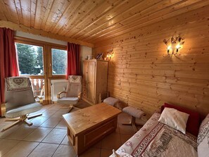 A cozy room with wooden walls, a wooden table, two chairs, a sofa, and a window with curtains. - 3-Room Apartment in Méribel - Sleeps 6 (Méribel)