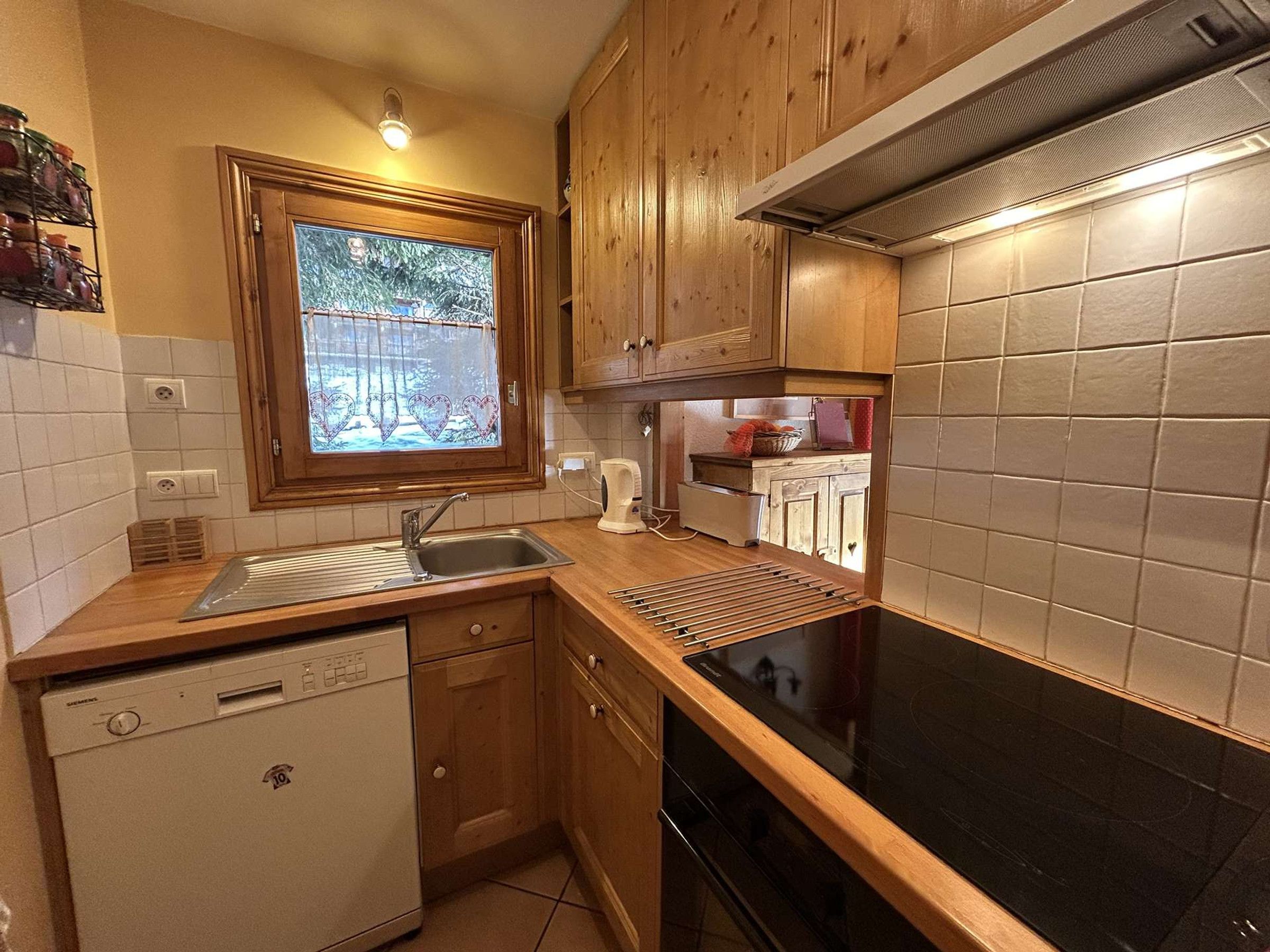 A compact kitchen with wooden cabinets, a built-in dishwasher, a stove, and a window with a view of trees.