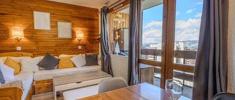 A cozy room with a wooden wall, a bed, a table with a glass, and a view of the city.