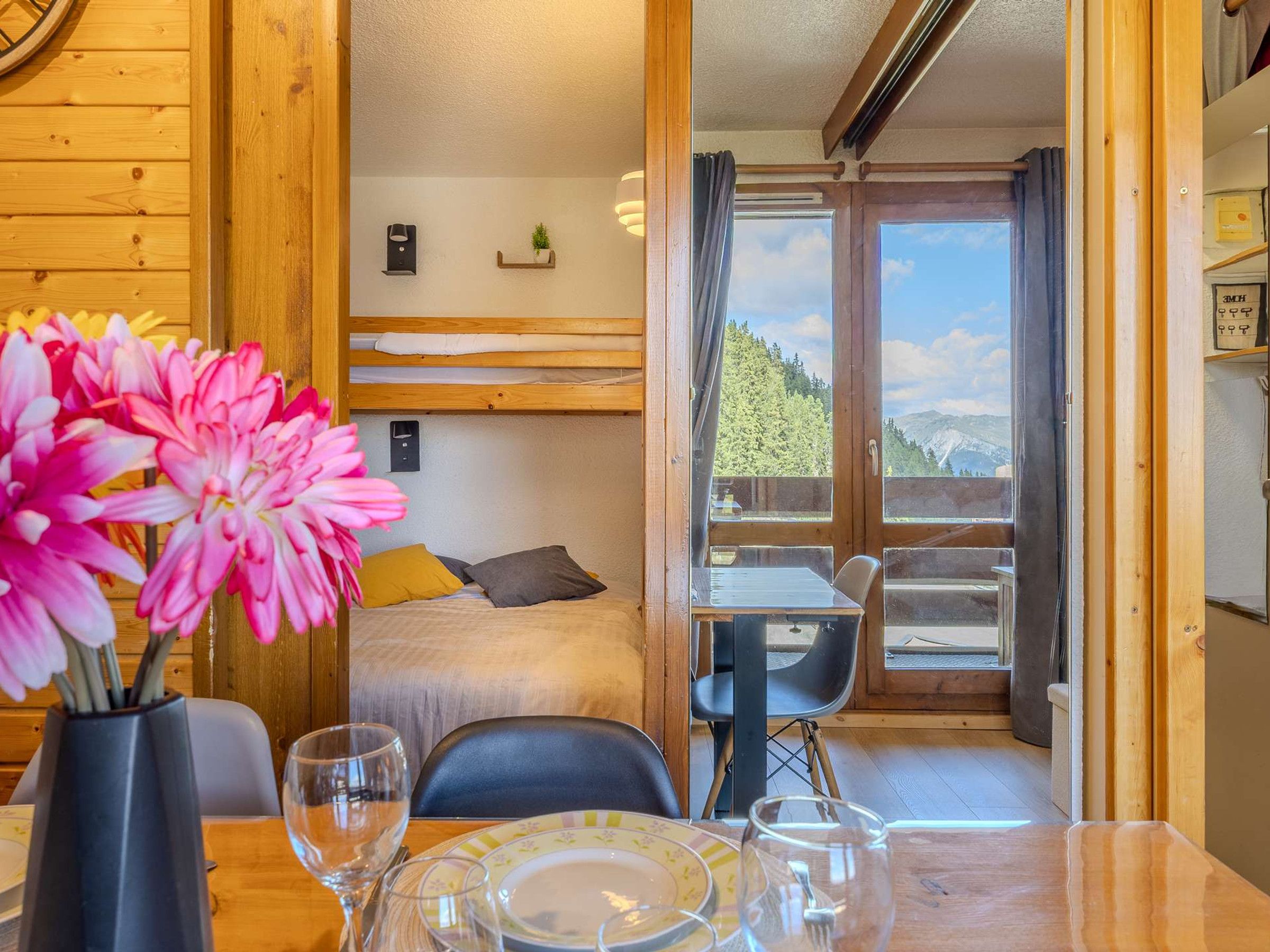 A room with a bed, a table with chairs, a window with a view of the outdoors, and a vase with flowers.
