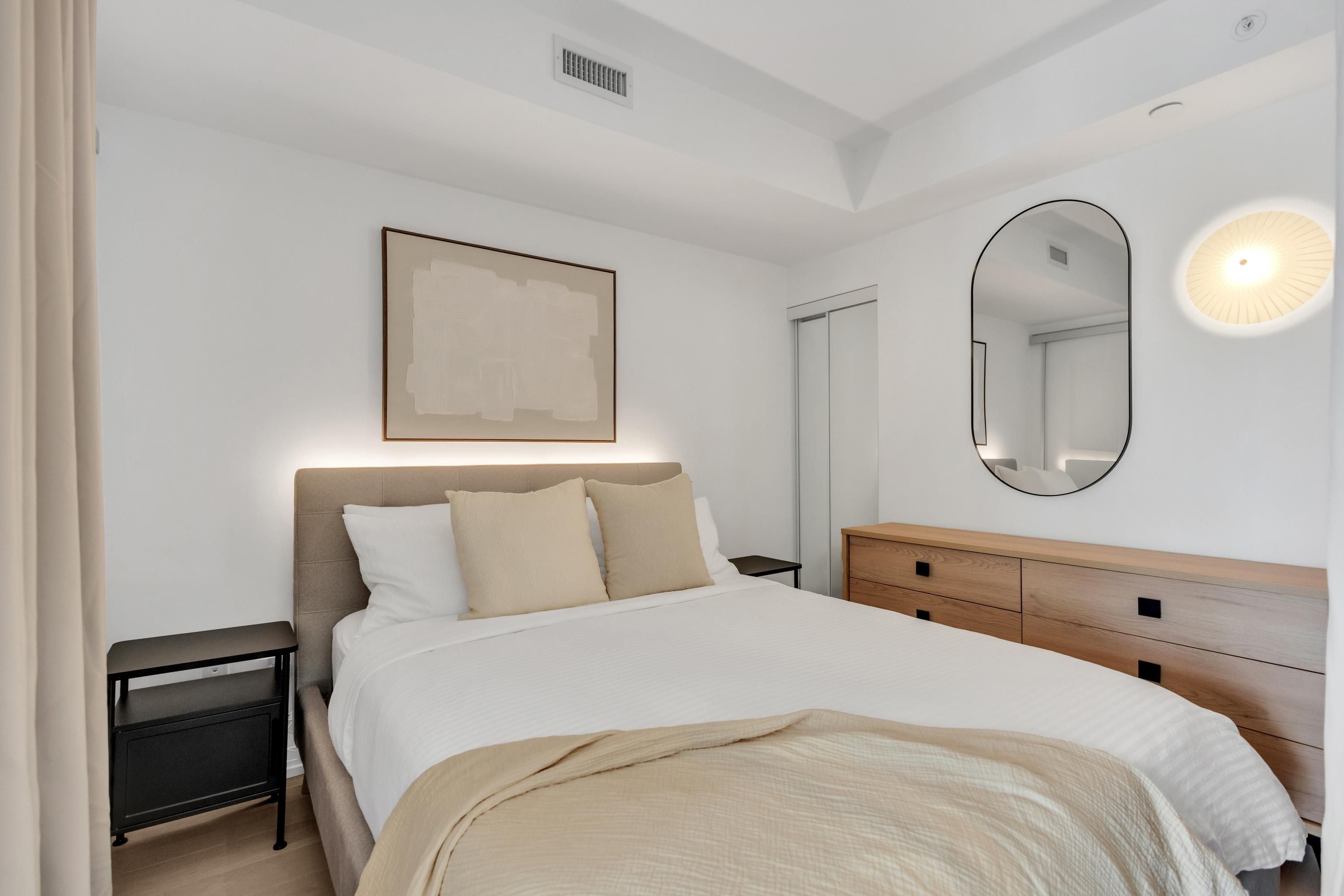 Un dormitorio con cama, mesitas de noche, cómoda, espejo y una obra de arte colgada en la pared.