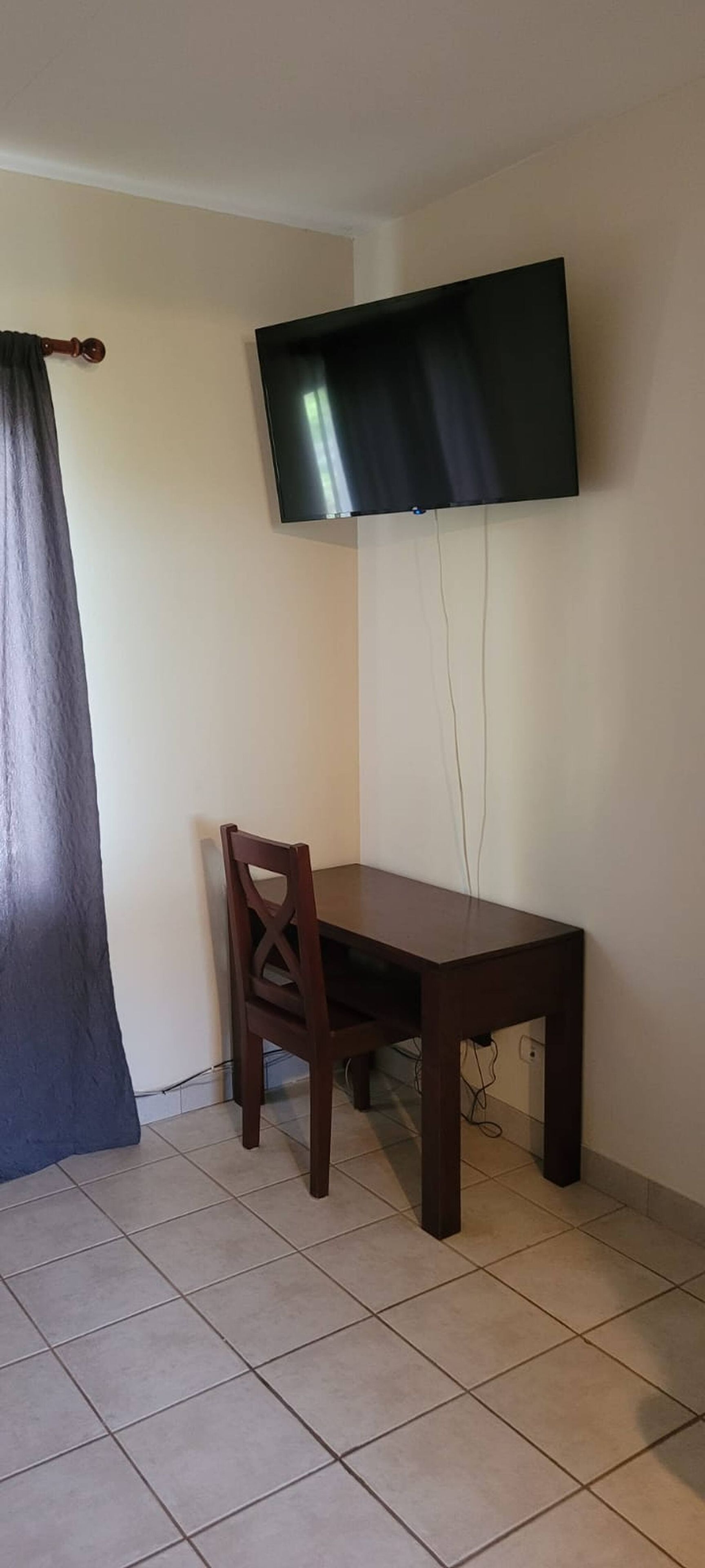 Una habitación con una televisión de pantalla plana montada en la pared, una mesa de comedor de madera, una silla y una cortina.