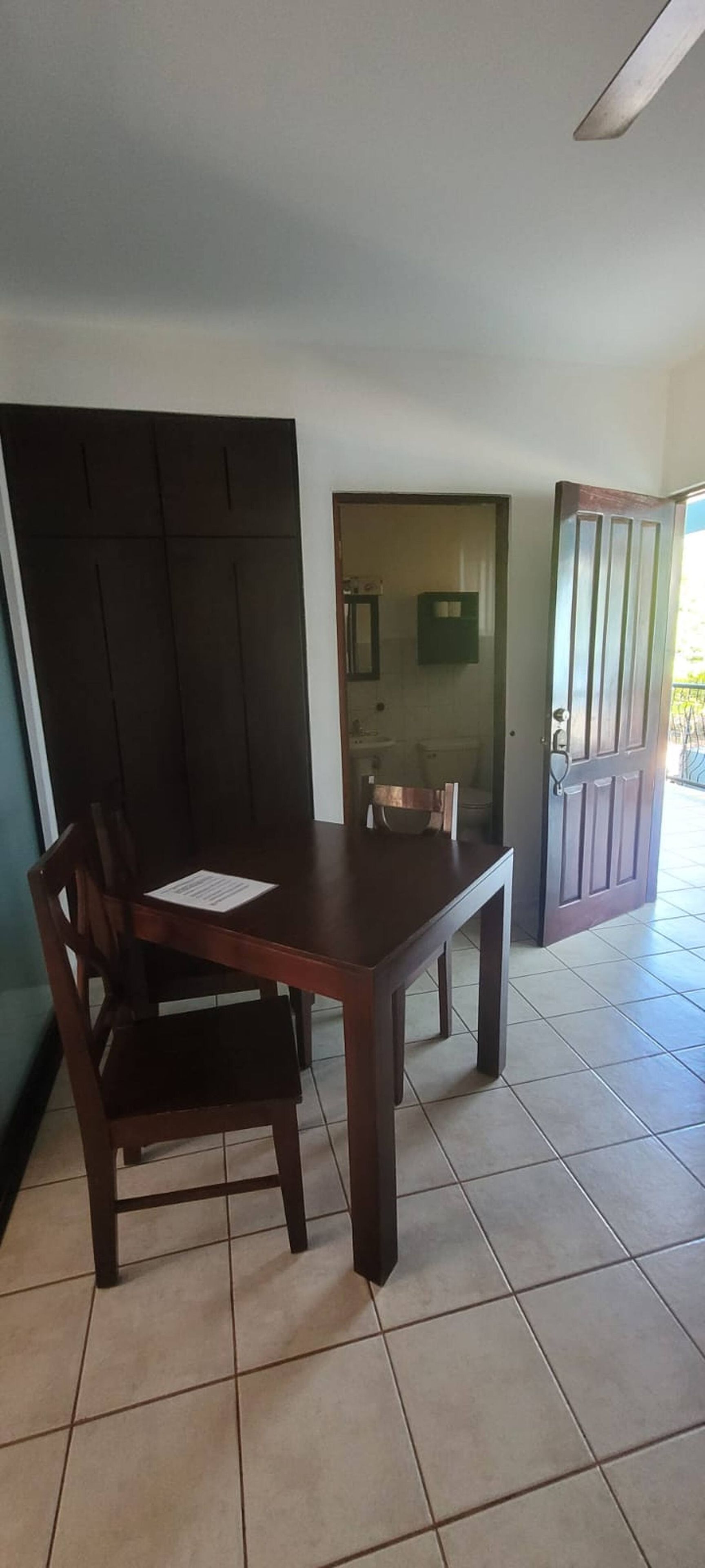 Zona de dinar amb taula i cadires, cuina a la vista per una porta oberta i un terra d'azulejos.