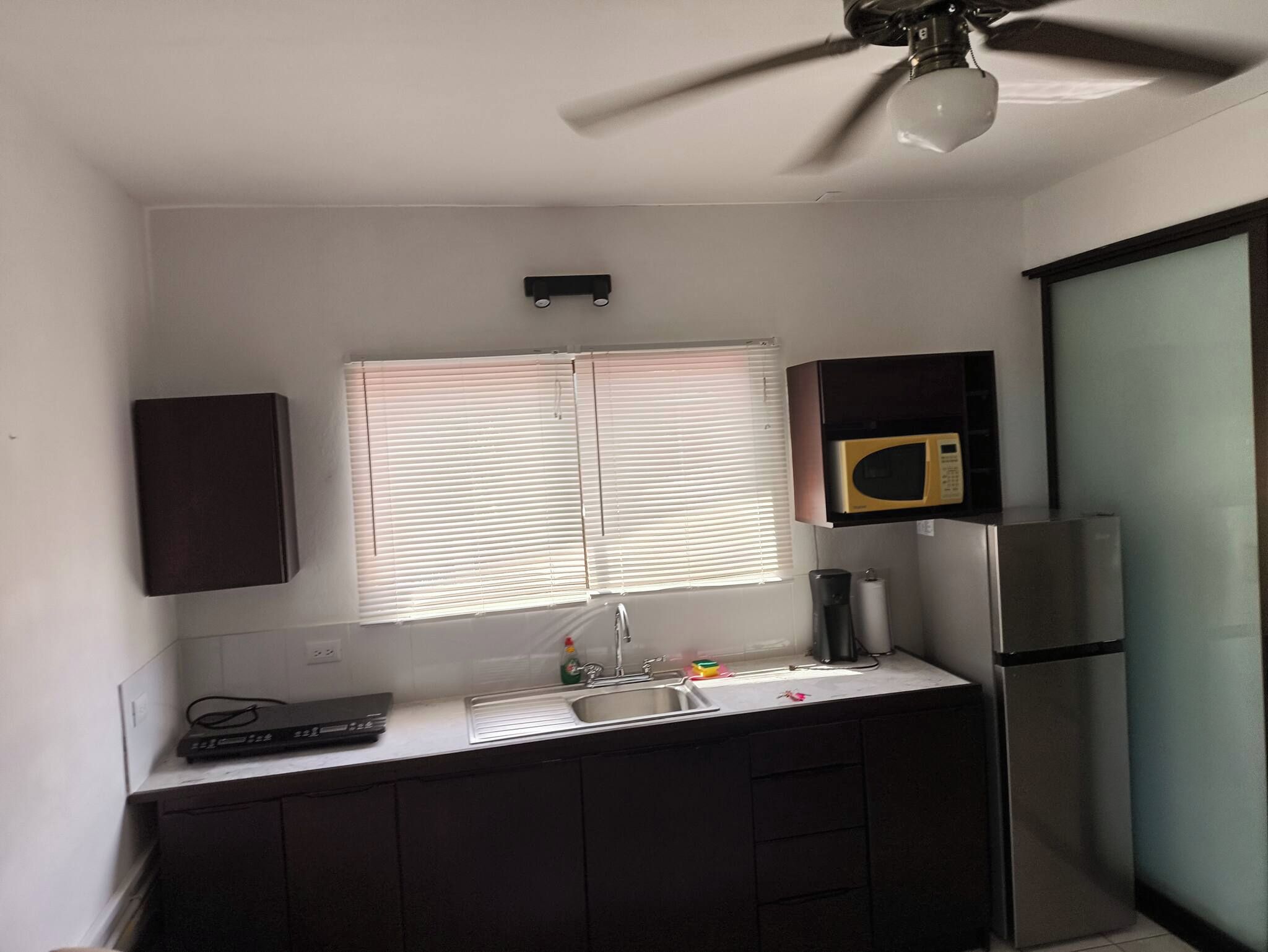 Cocina compacta con microondas integrado, refrigerador de acero inoxidable y ventana con persianas.