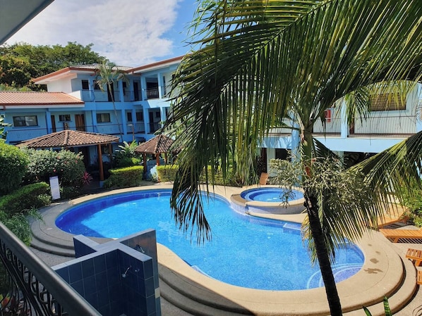 Piscina com hidromassagem, rodeada por palmeiras e um edifício com varandas.