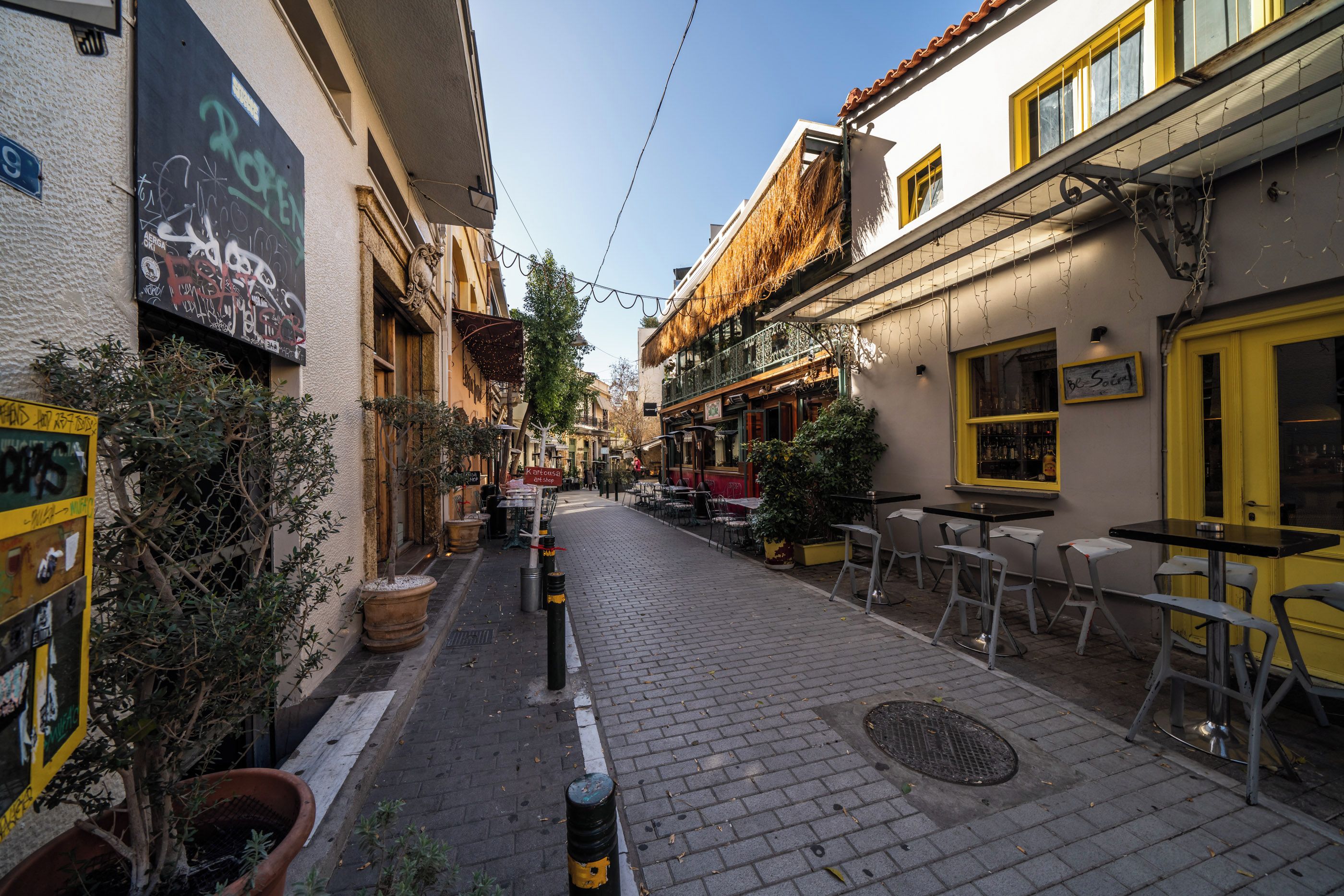 A narrow street with buildings on either side, outdoor seating, and a sign for a shop.