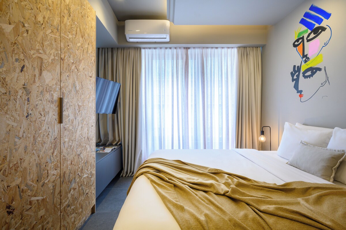 A hotel room with a bed, a television, a wall-mounted air conditioner, and a window with curtains.