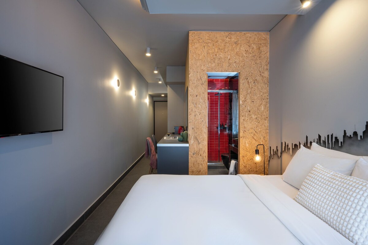 A modern hotel room with a large bed, a flat-screen TV mounted on the wall, and a cityscape silhouette on the wall.