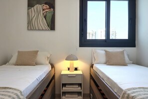 A bedroom with two single beds, a nightstand with a lamp, a painting on the wall, and a window with a view of buildings. - SUNSET WAVES elegant apartment (Benidorm)
