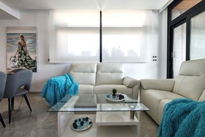 Un salon moderne avec une table basse en verre, un canapé beige et un tableau représentant une femme au bord de la mer.