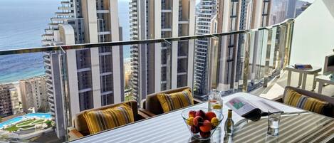 A balcony with a glass table, cushions, and a view of tall buildings and the sea.