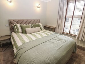 A bedroom with a bed, bedside tables, a nightstand, a window with blinds, and a wall-mounted light fixture. - Corner House (Hexham)
