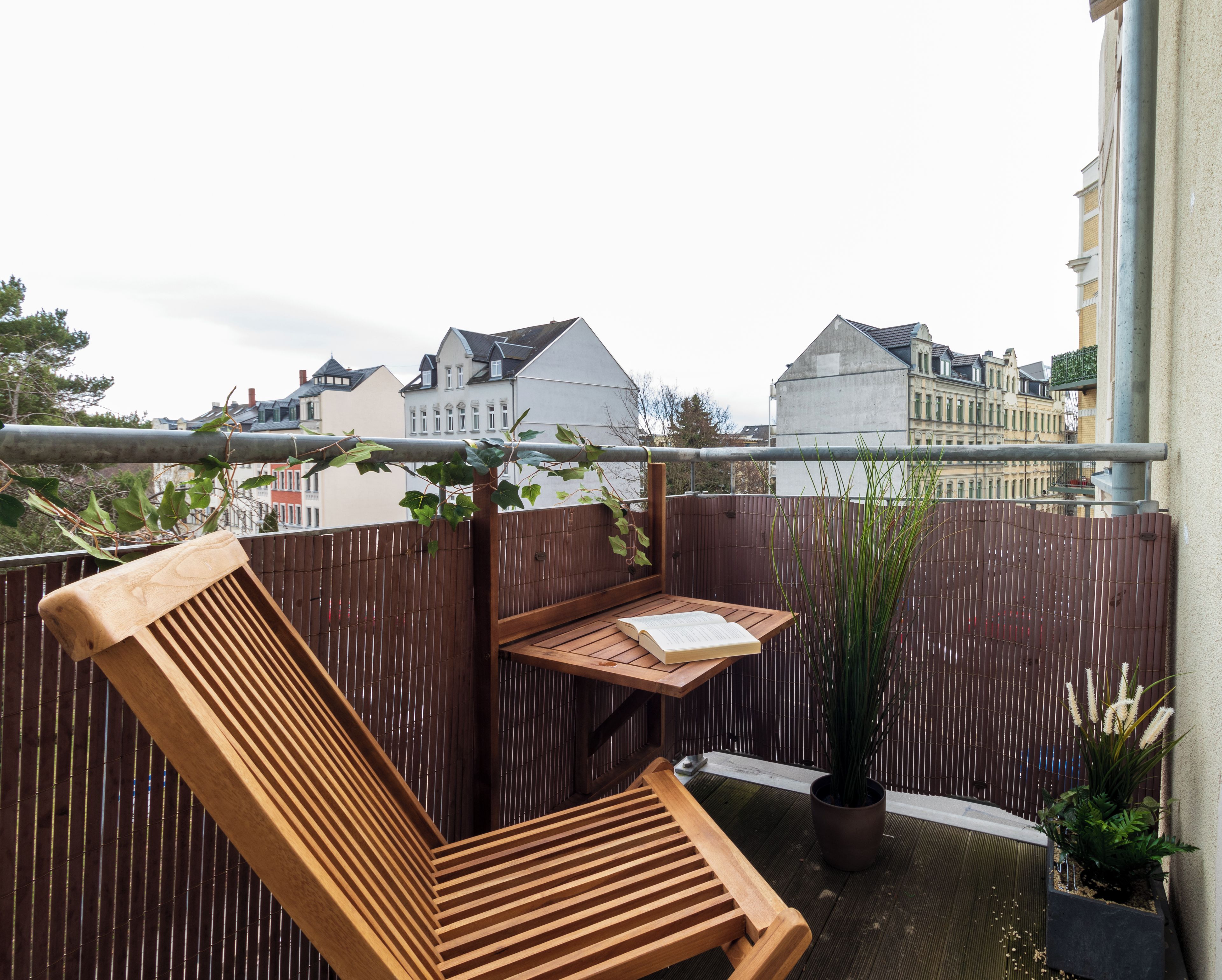 Eine Holzbank mit einem Buch, einer Topfpflanze und Blick auf Gebäude im Hintergrund.