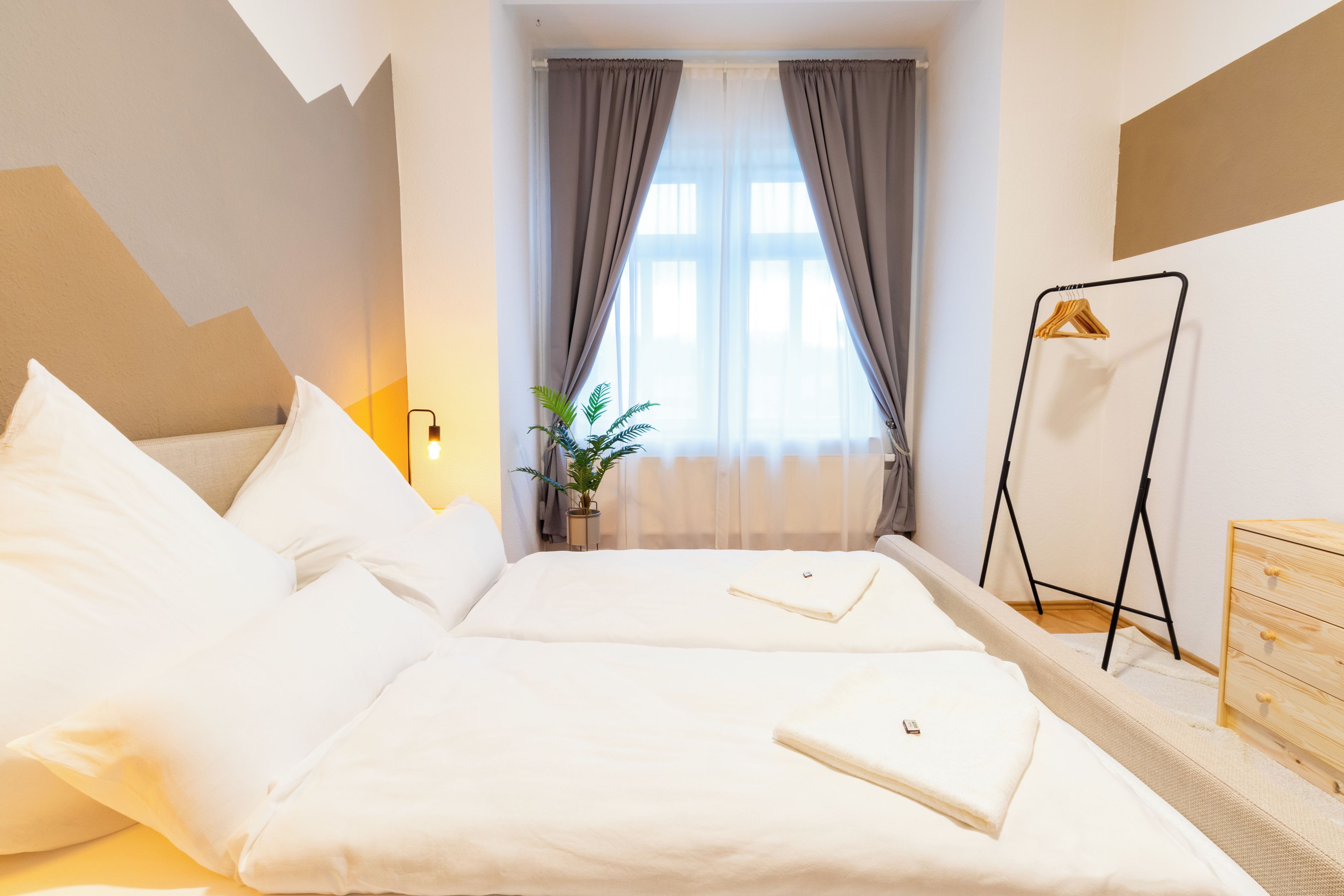 Ein Hotelzimmer mit Bett, Nachttisch, Pendelleuchte, Topfpflanze und Kleiderhaken.
