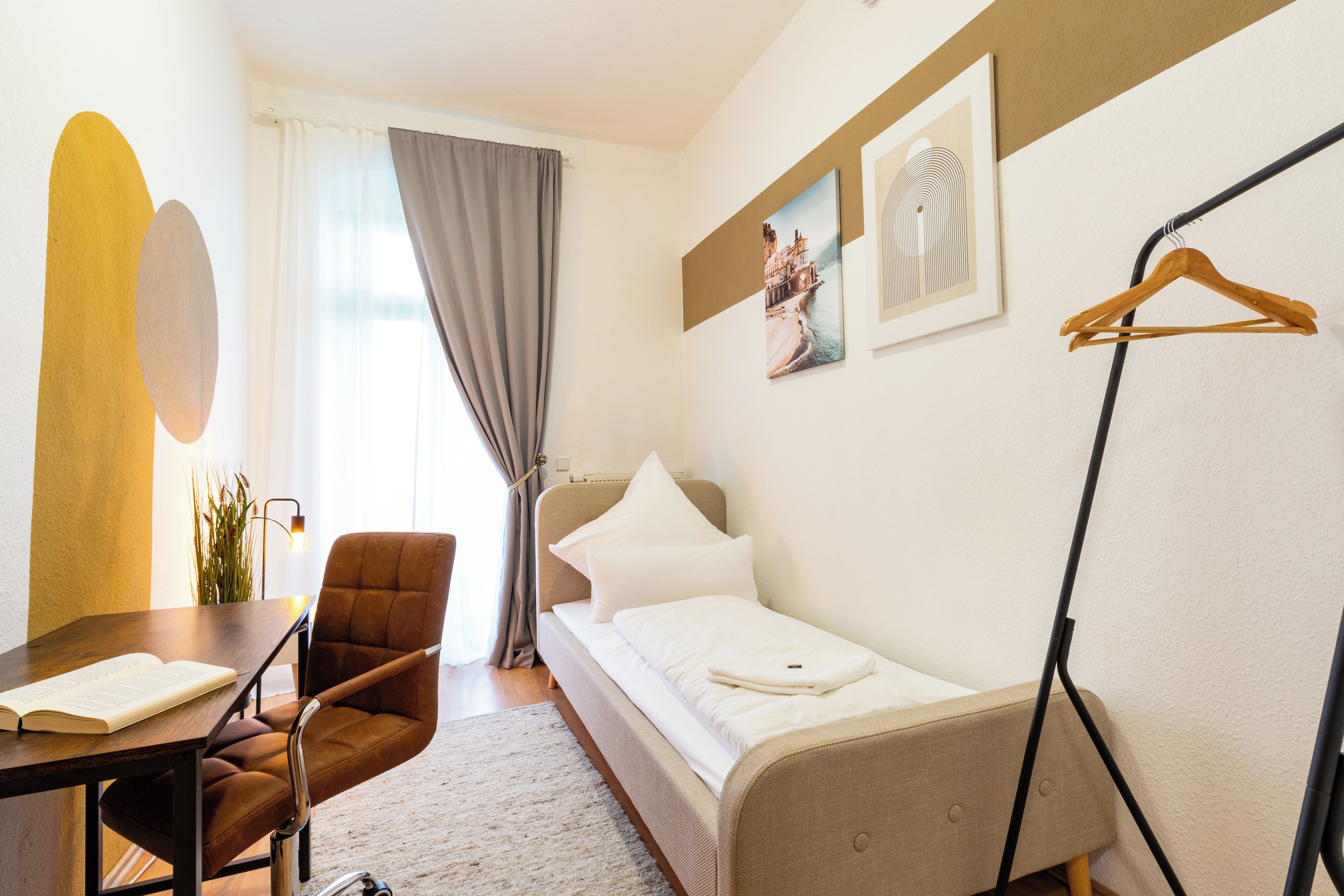 Ein Hotelzimmer mit Bett, einem Sessel, einem Tisch mit einem Buch, einem Kleiderhaken und einem Fenster mit Vorhängen.