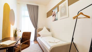Ein Hotelzimmer mit Bett, einem Sessel, einem Tisch mit einem Buch, einem Kleiderhaken und einem Fenster mit Vorhängen.