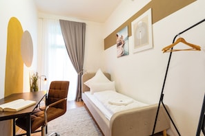 A hotel room with a bed, a chair, a table with a book, a hanging coat rack, and a window with curtains.