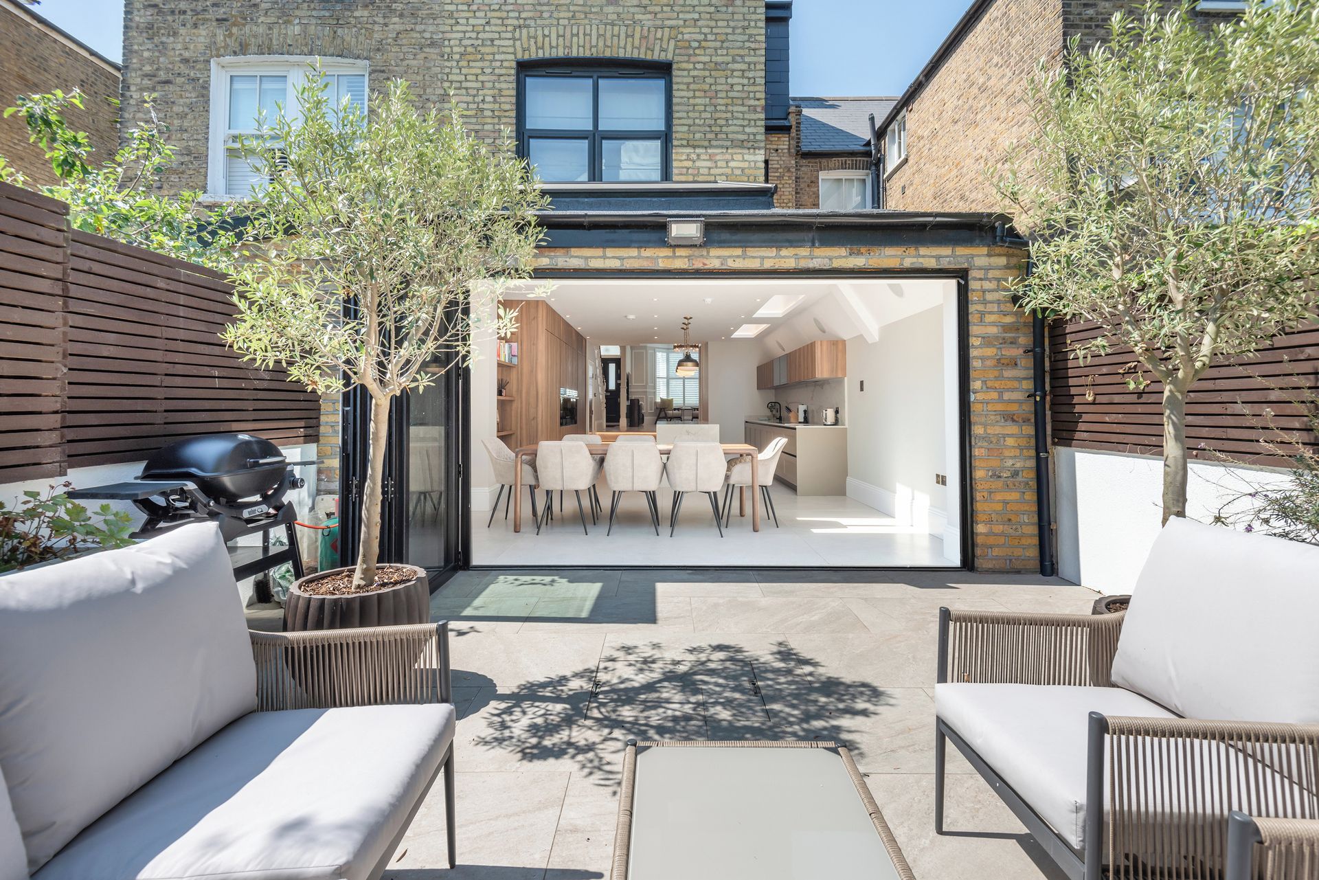 A modern patio with outdoor furniture, a barbecue grill, and a glass door leading to a kitchen and dining area.