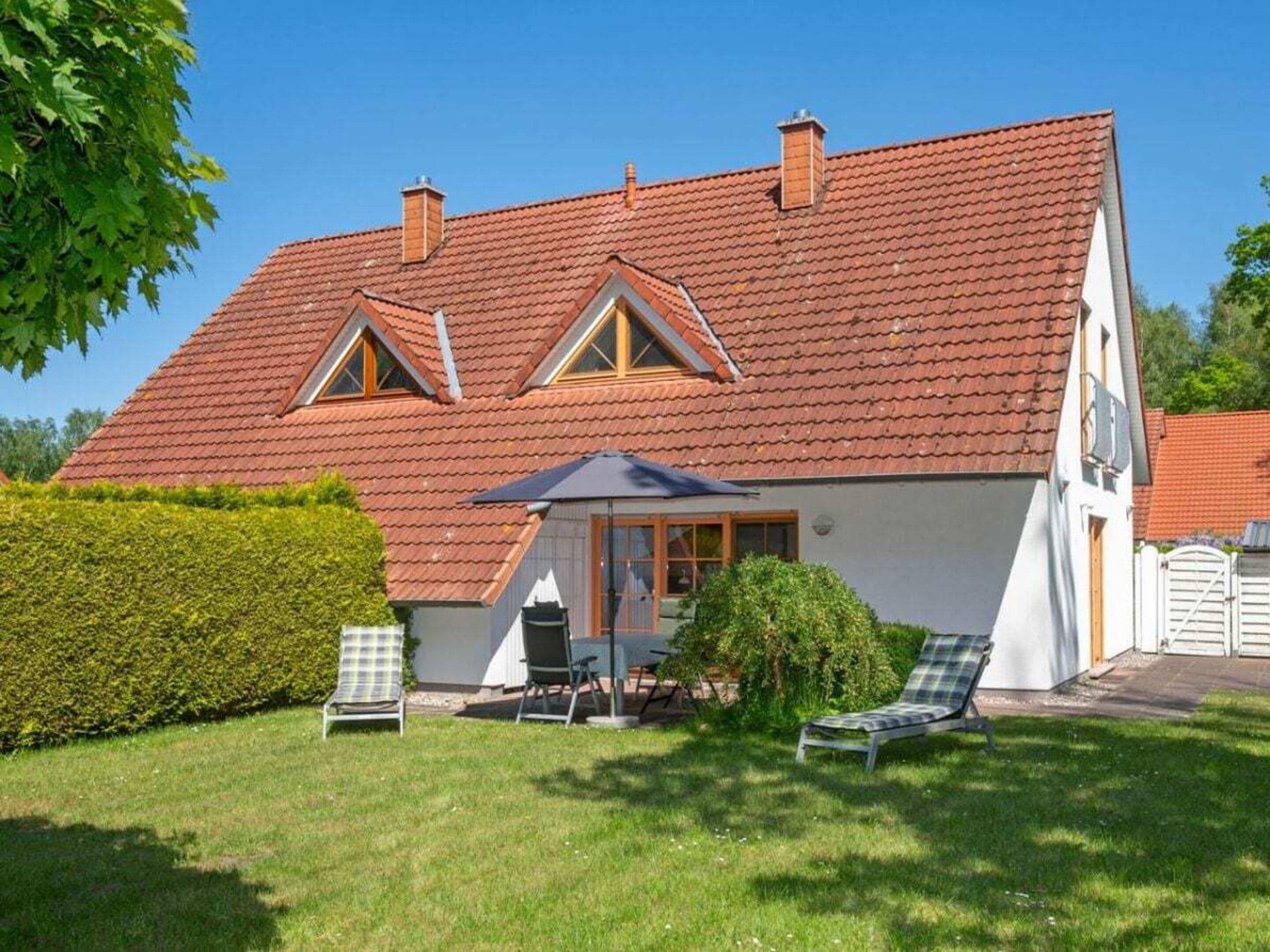 A two-story house with a red-tiled roof, a covered patio with a table and chairs, and a lawn with a bench.