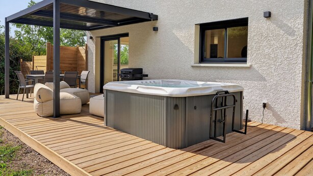 Een hottub op een houten terras, compleet met een pergola erboven, een patio tafel met stoelen en een raam in het huis.