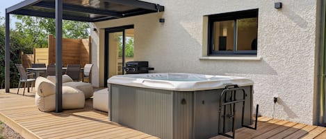 Ein Whirlpool auf einer Holzterrasse mit einer Pergola darüber, ein Patio-Tisch mit Stühlen und ein Fenster am Haus.