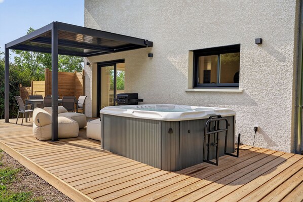 Uma banheira de hidromassagem em um deck de madeira, com uma pérgola acima, uma mesa de pátio com cadeiras e uma janela na casa.