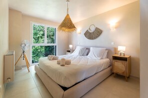 Um quarto espaçoso com uma cama grande, duas mesas de cabeceira com luminárias, um ventilador e vista para o verde pela janela.