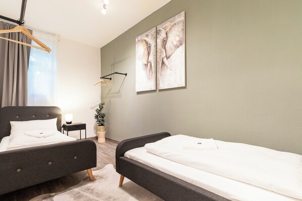 A hotel room with two beds, a nightstand, a lamp, a plant, and a painting on the wall.