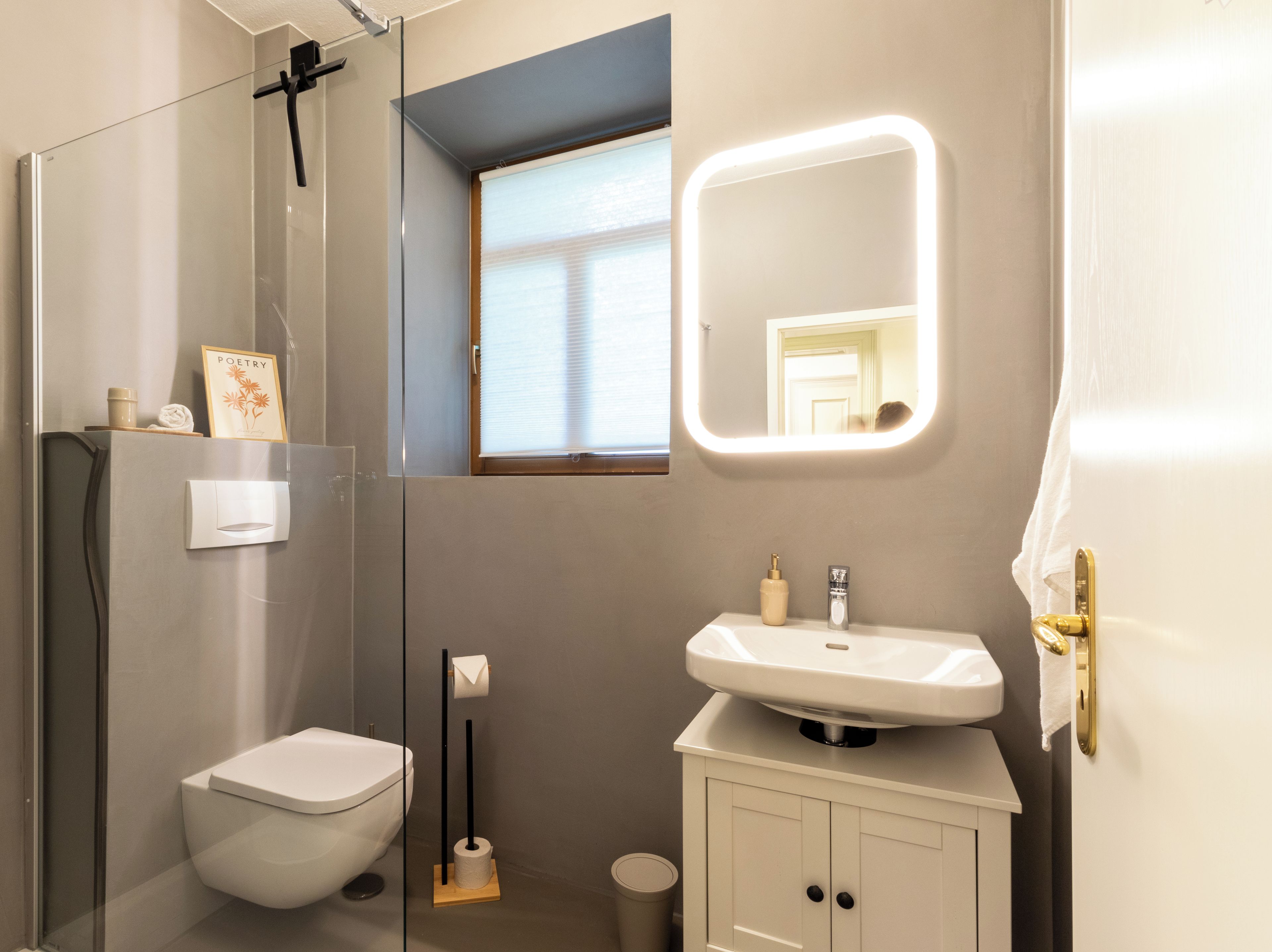 Ein modernes Badezimmer mit Toilette, Waschbecken, einem beleuchteten Spiegel, einem Fenster und einem Handtuchhalter.