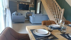 A dining area with a table set for two, a sofa, and a staircase in the background. - Villa 'Piscine 9pers SaôneBeaujolais' with Private Pool, Shared Terrace and Wi-Fi (Montmerle-sur-Saône)