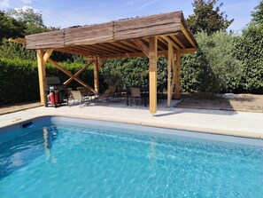 A pool area with a wooden pergola, lounge chairs, and a barbecue grill.