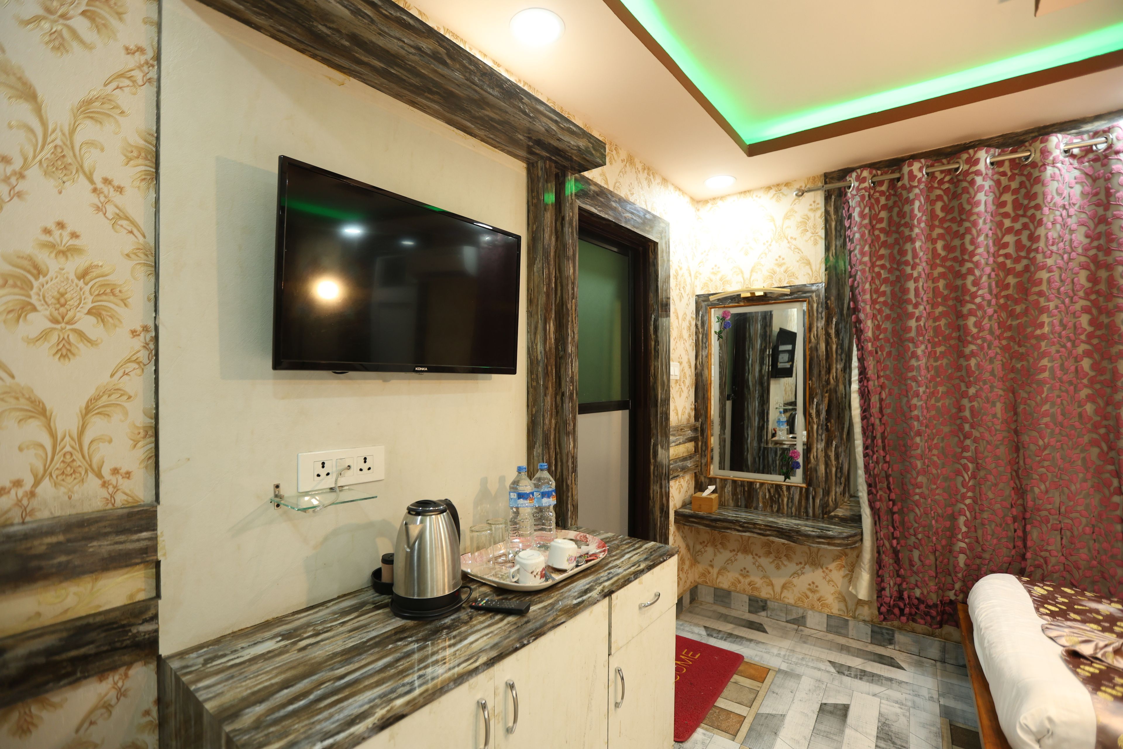 Quarto de hotel com televisão de tela plana na parede, cafeteira no balcão, espelho e cama com tapete vermelho.