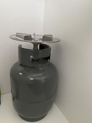Un cilindro de gas grande, gris, con una parte superior metálica y un asa, apoyado contra una pared lisa.