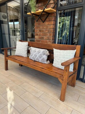 A wooden bench with patterned cushions in front of a brick wall and glass doors. - Exquisite Art Deco 2 bed Apartment - 2395 (Harare)