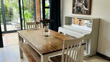 A dining area with a wooden table and white chairs, a piano, and a large framed artwork on the wall.