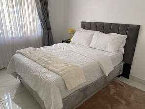 Ein ordentlich bezogenes Bett mit grauem Kopfteil, weißer Bettwäsche und einem braunen Teppich.
