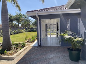 A house with a gated entrance, a paved driveway, and a well-maintained garden with palm trees and other plants. - Newly Renovated 4 Bedroom Guesthouse - 2389 (Harare)