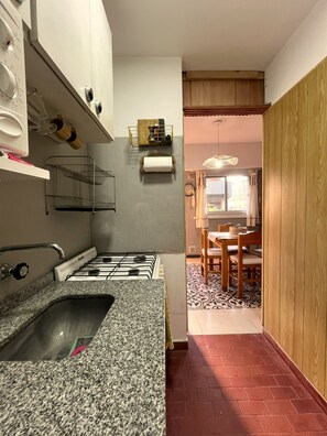 A compact kitchen with a granite countertop, a gas stove, and a wooden dining area with a patterned floor. - Dept. "Caracolas" | Half a Block from the Beach - Ayres del Bosque Lodging (Valeria del Mar)