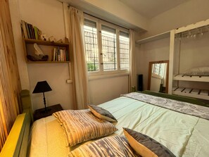 A bedroom with a bed, pillows, a bedside table with a lamp, a shelf with books, and a window with curtains. - Dept. "Caracolas" | Half a Block from the Beach - Ayres del Bosque Lodging (Valeria del Mar)