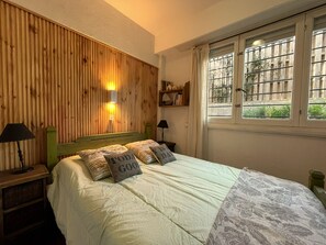Ein Schlafzimmer mit hölzernem Kopfteil, einem Bett mit gemusterten Kissen, einem Nachttisch mit Lampe und einem Fenster mit Blick auf Grünflächen.