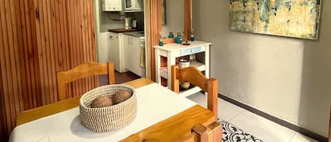 Un comedor con mesa y sillas de madera, una cesta tejida, una lámpara colgante moderna, una cocina al fondo y un cuadro abstracto de gran tamaño en la pared.