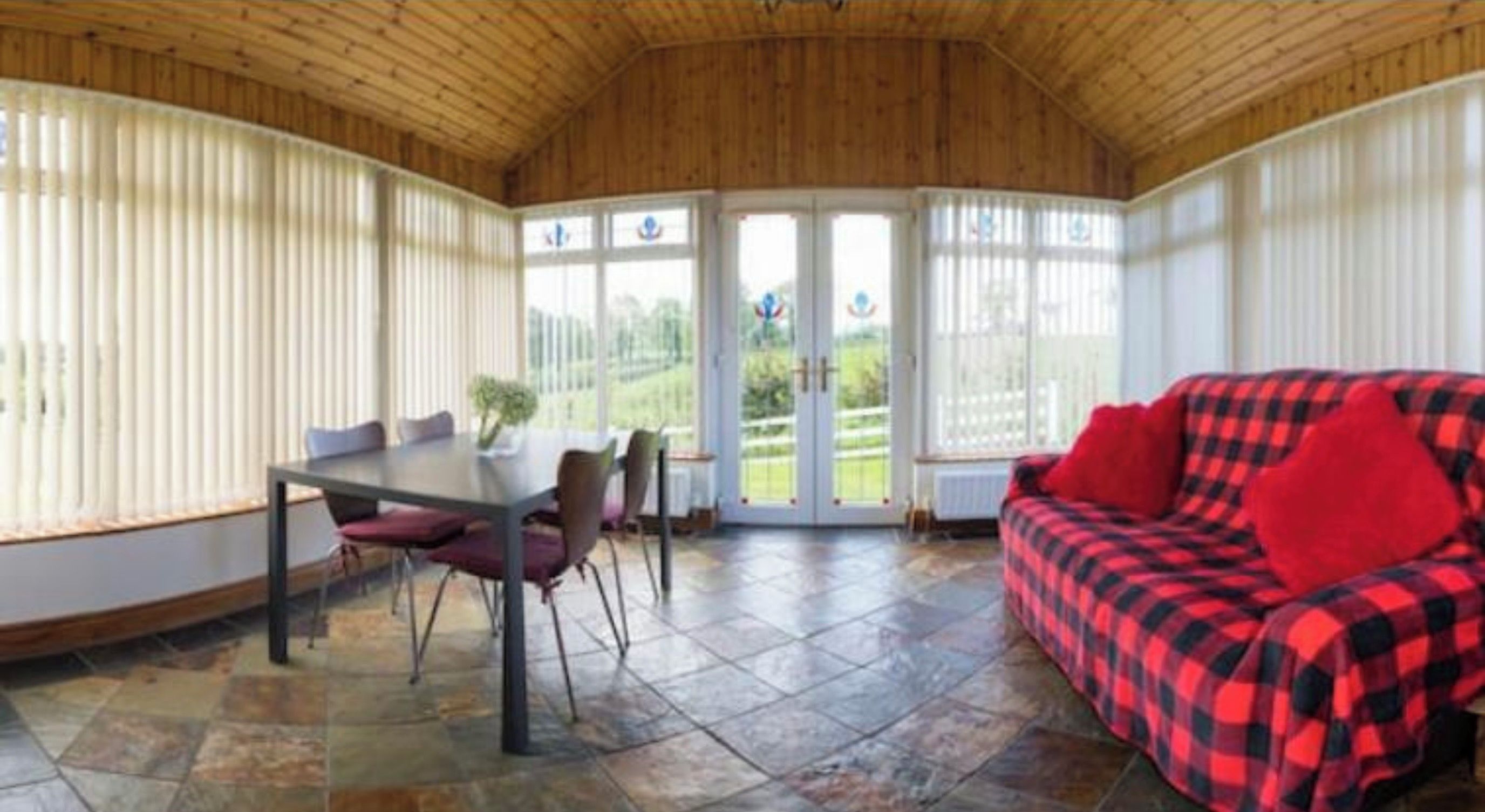 Una habitación con una mesa de comedor y sillas, un sofá rojo y negro a cuadros, y vistas a través de un ventanal.