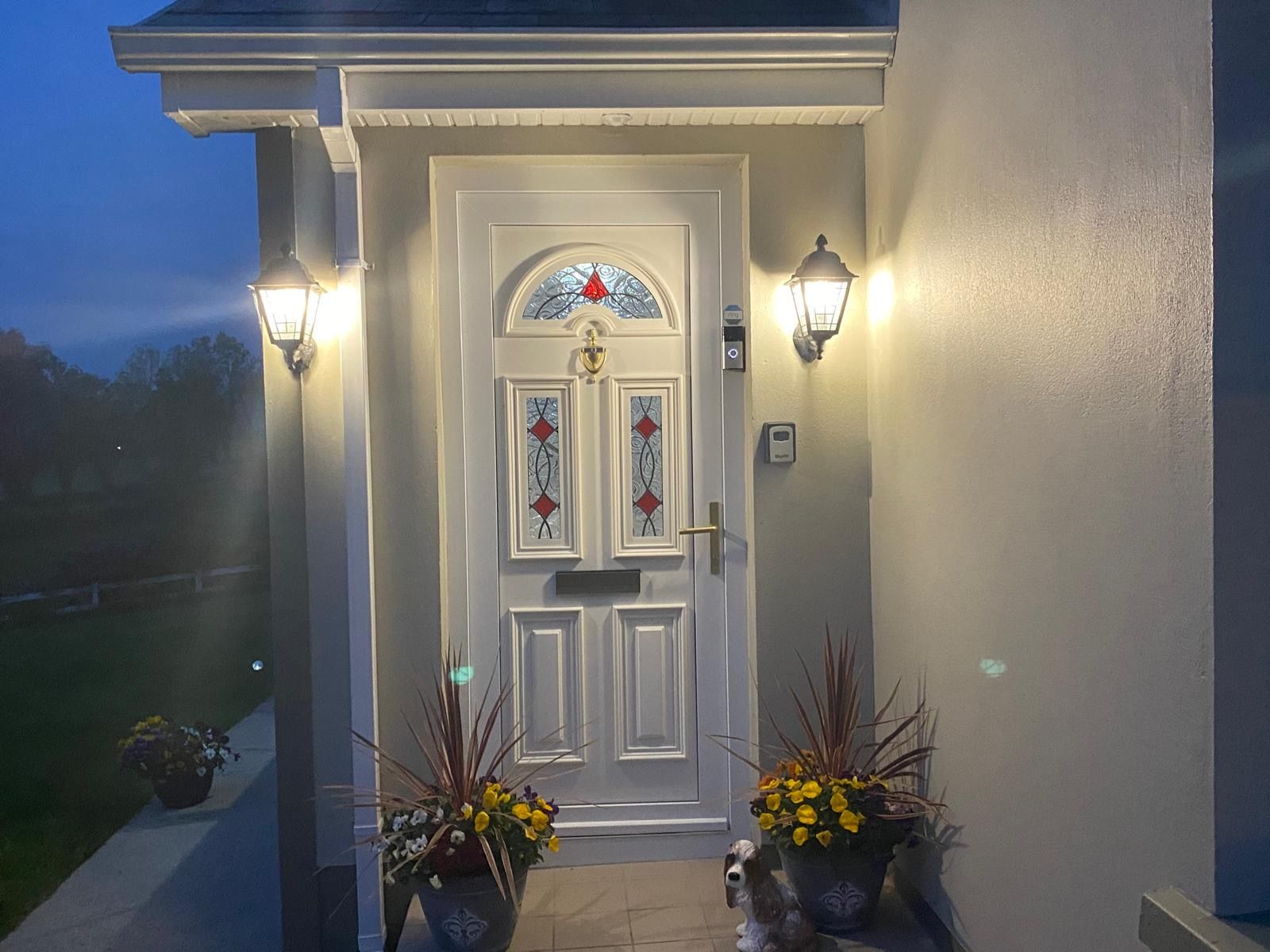 A white door with a decorative glass panel, two wall-mounted lights, and two potted plants on the porch.