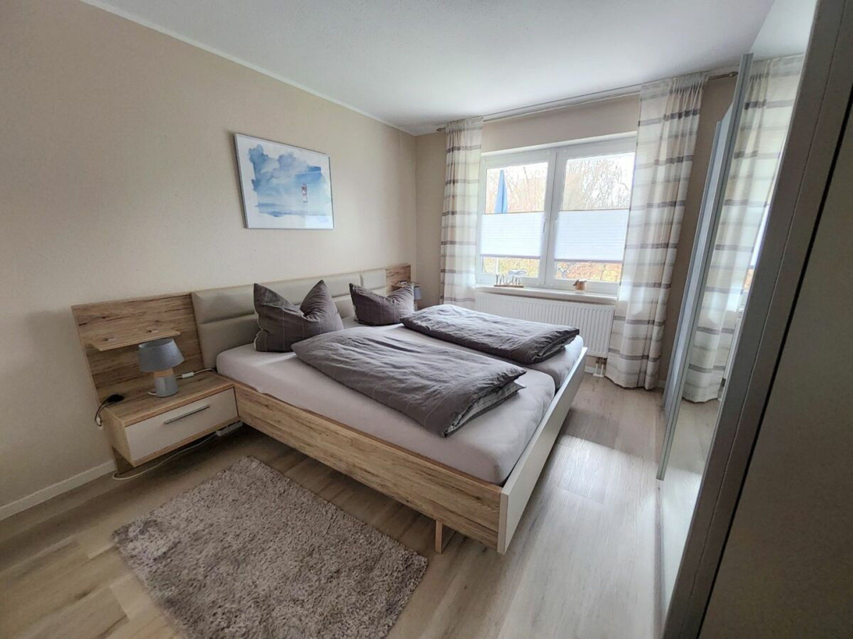 Un dormitorio con cama, mesitas de noche, lámpara, ventana con cortinas y un cuadro en la pared.