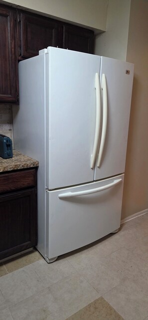 A white refrigerator with two doors, a built-in ice maker, and a freezer drawer, located in a kitchen with dark wooden cabinets and a tiled backsplash.