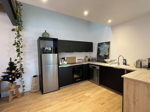A modern kitchen with a built-in oven, microwave, and refrigerator. There is a wooden dining table and a small wooden stool. A Christmas tree with lights is visible in the corner. - Bruisse Vacation Rental (Saint-Brieuc-des-Iffs)