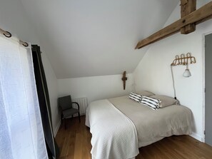 A bedroom with a sloped ceiling, a bed with a white quilt, a patterned chair, and a wall-mounted lamp. - Bruisse Vacation Rental (Saint-Brieuc-des-Iffs)
