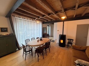 A dining area with a round table and chairs, a sofa, a fireplace, and a view through a window with sheer curtains. - Bruisse Vacation Rental (Saint-Brieuc-des-Iffs)